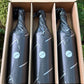 Three bottles wrapped in black paper with a blue label in a cardboard box outdoors.