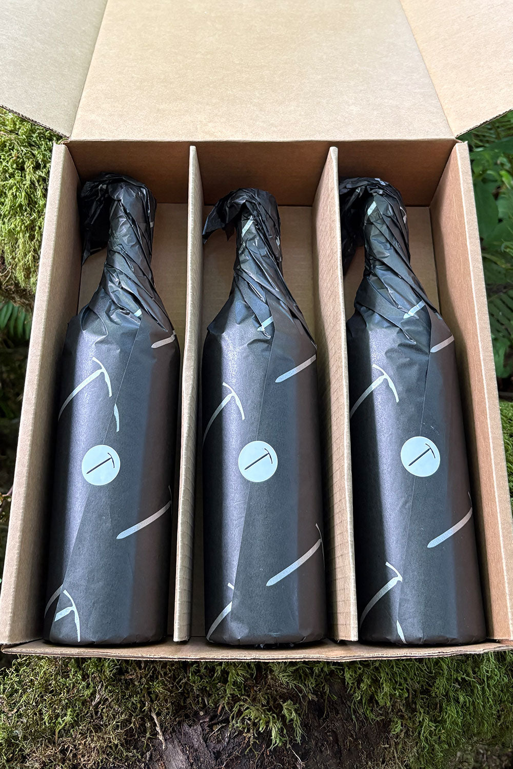 Three bottles wrapped in black paper with a blue label in a cardboard box outdoors.